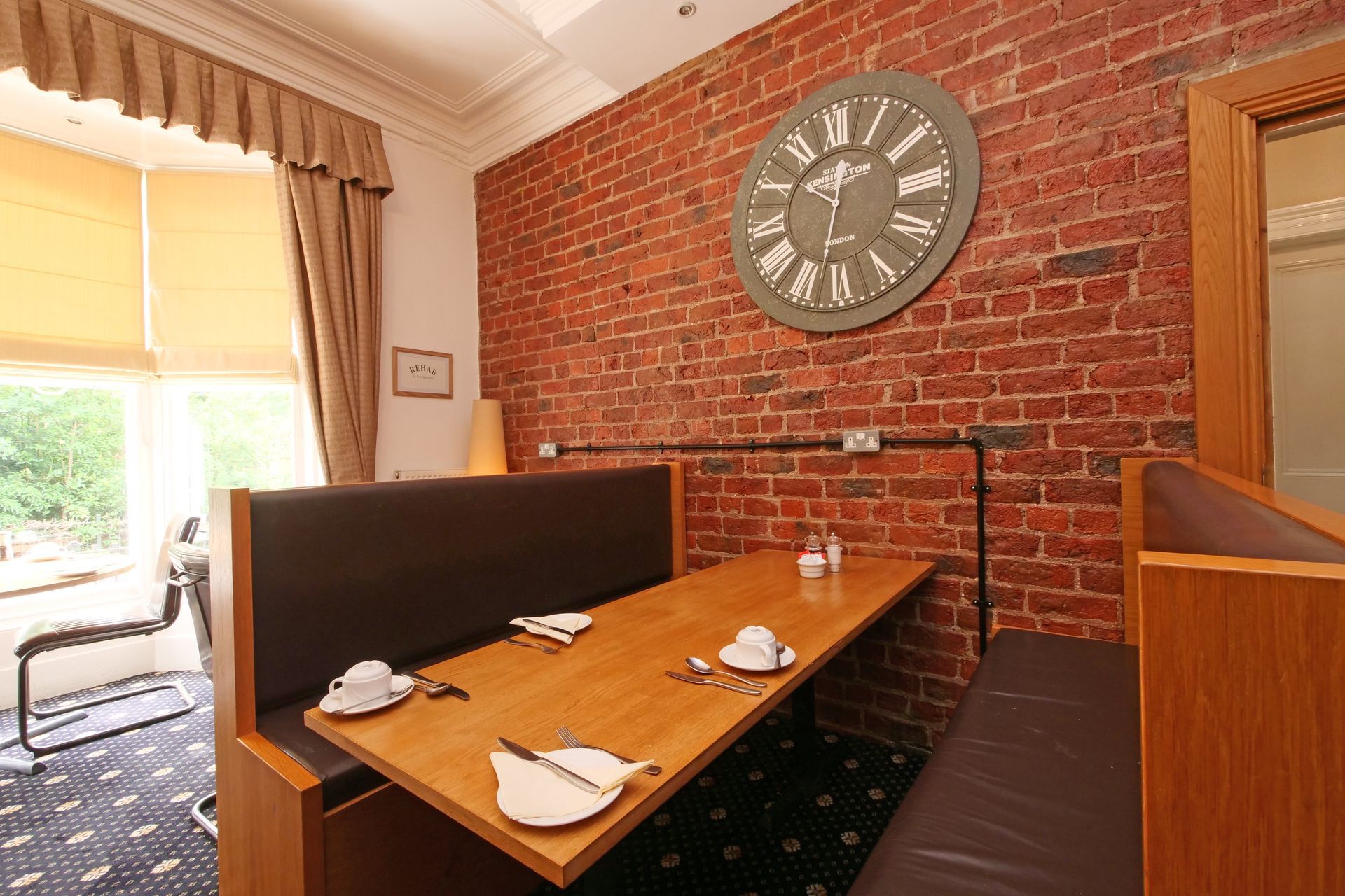 Restaurant booth with red brick wall, large clock, and wooden table set with dishes.