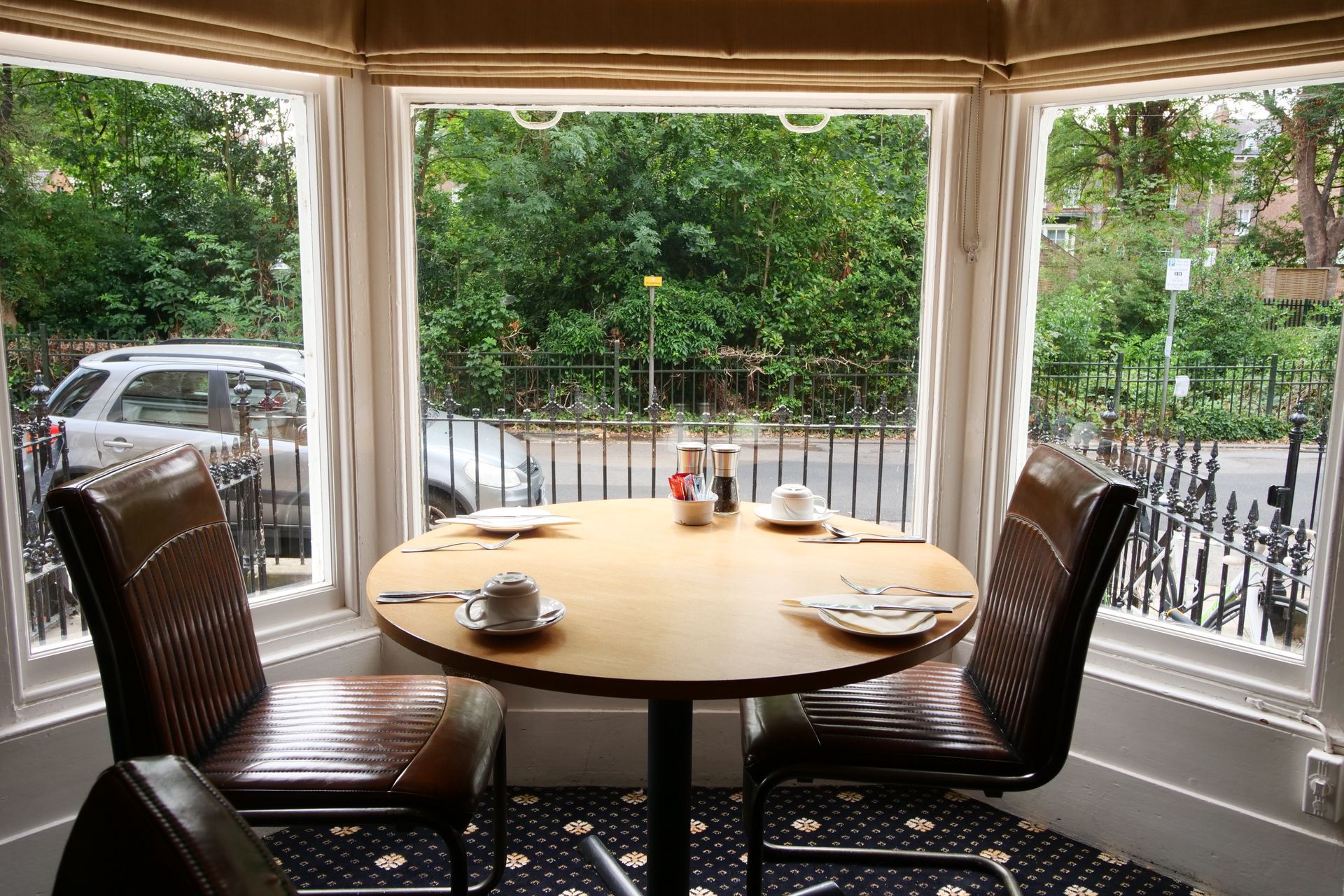 Table set for two in a bay window, with a view of a street; two chairs, a car, and trees.