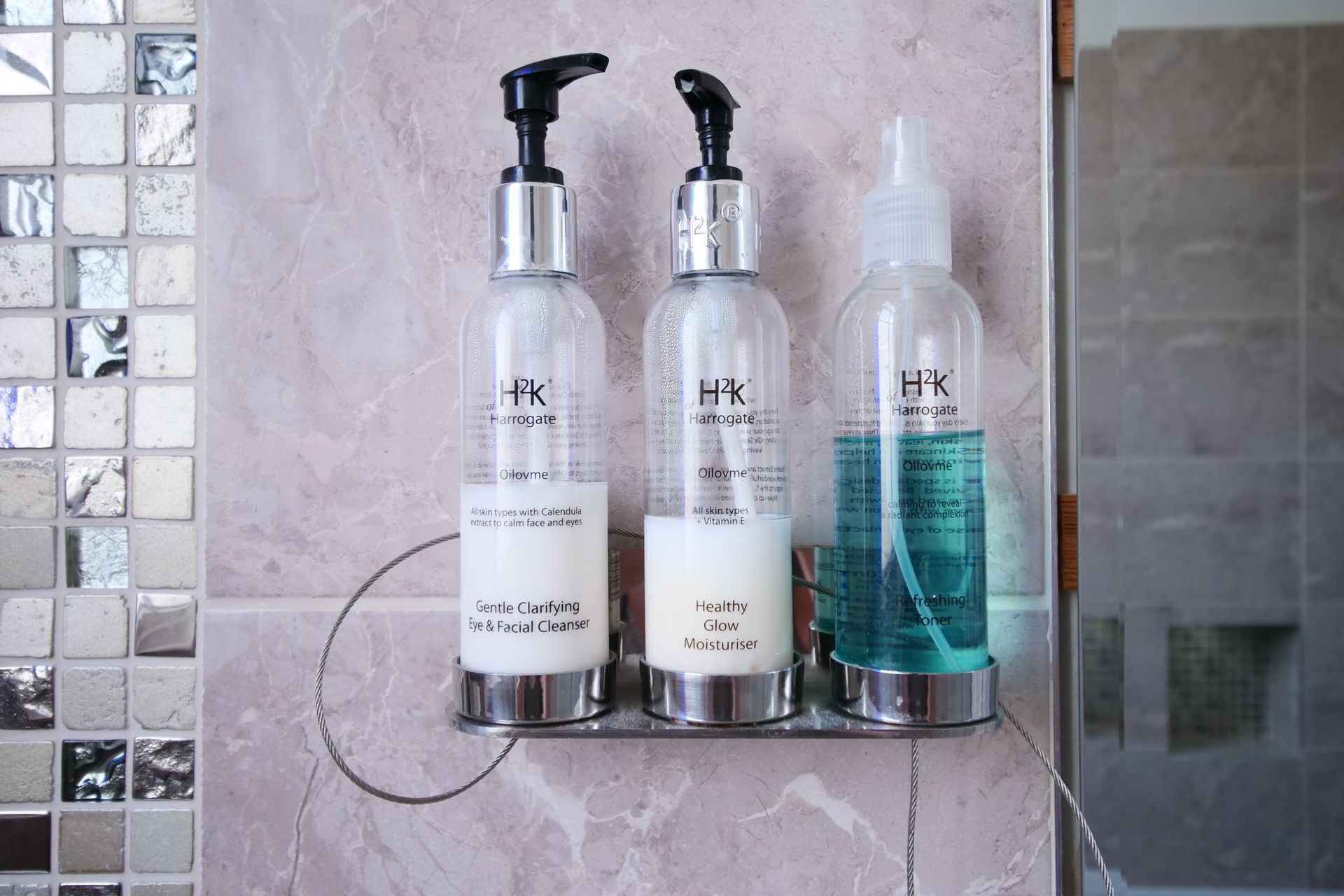 Three clear bottles of toiletries in a shower, mounted on a wall, featuring body wash, lotion, and spray.