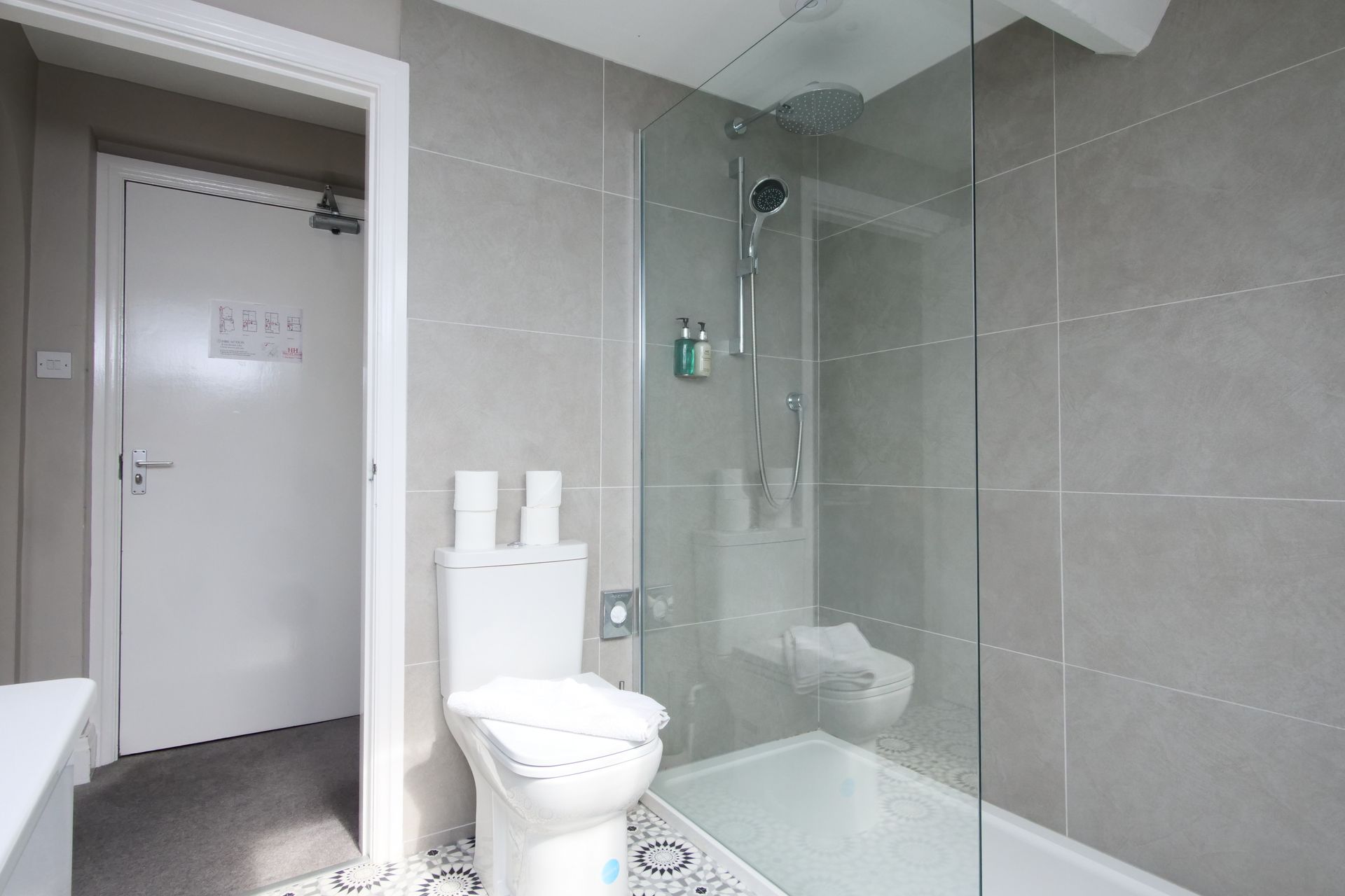 Bathroom with toilet, shower, and door; gray tiled walls, white fixtures.