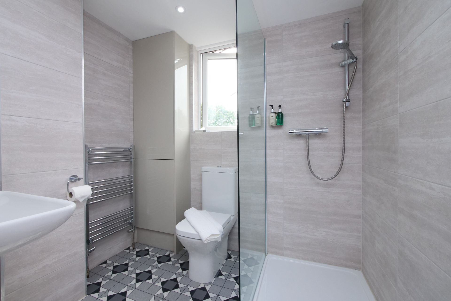 Bathroom with a toilet, sink, shower, and heated towel rack. Light gray walls, patterned floor.