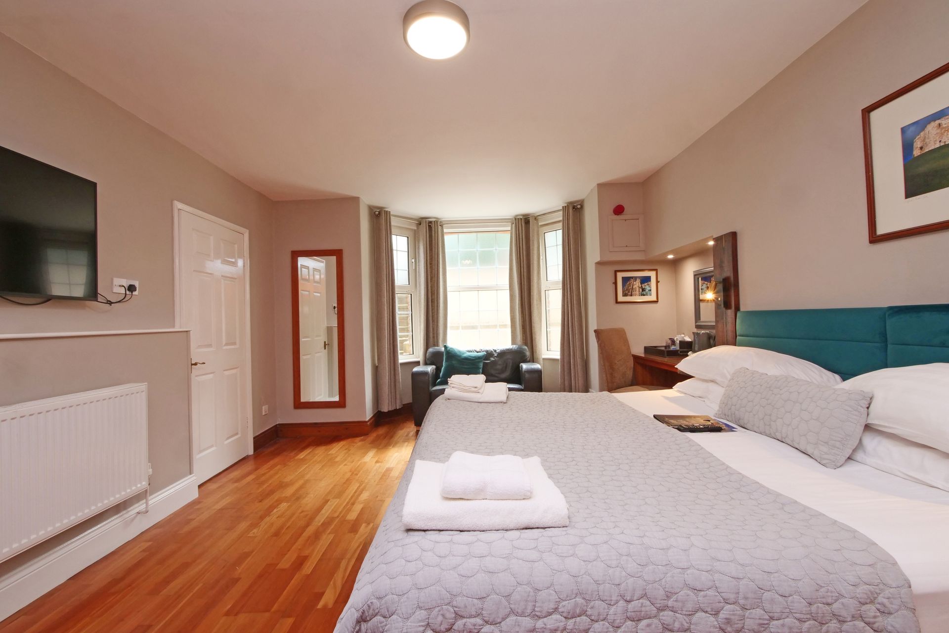 Hotel bedroom with double bed, TV, and bay window. Wooden floor, gray walls, turquoise headboard.