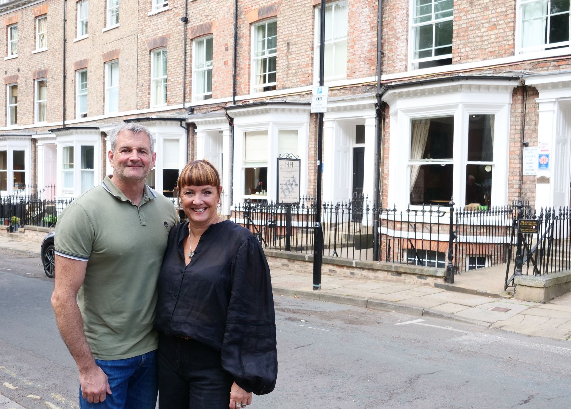 Man and woman posing on street in front of brick building with white bay windows.