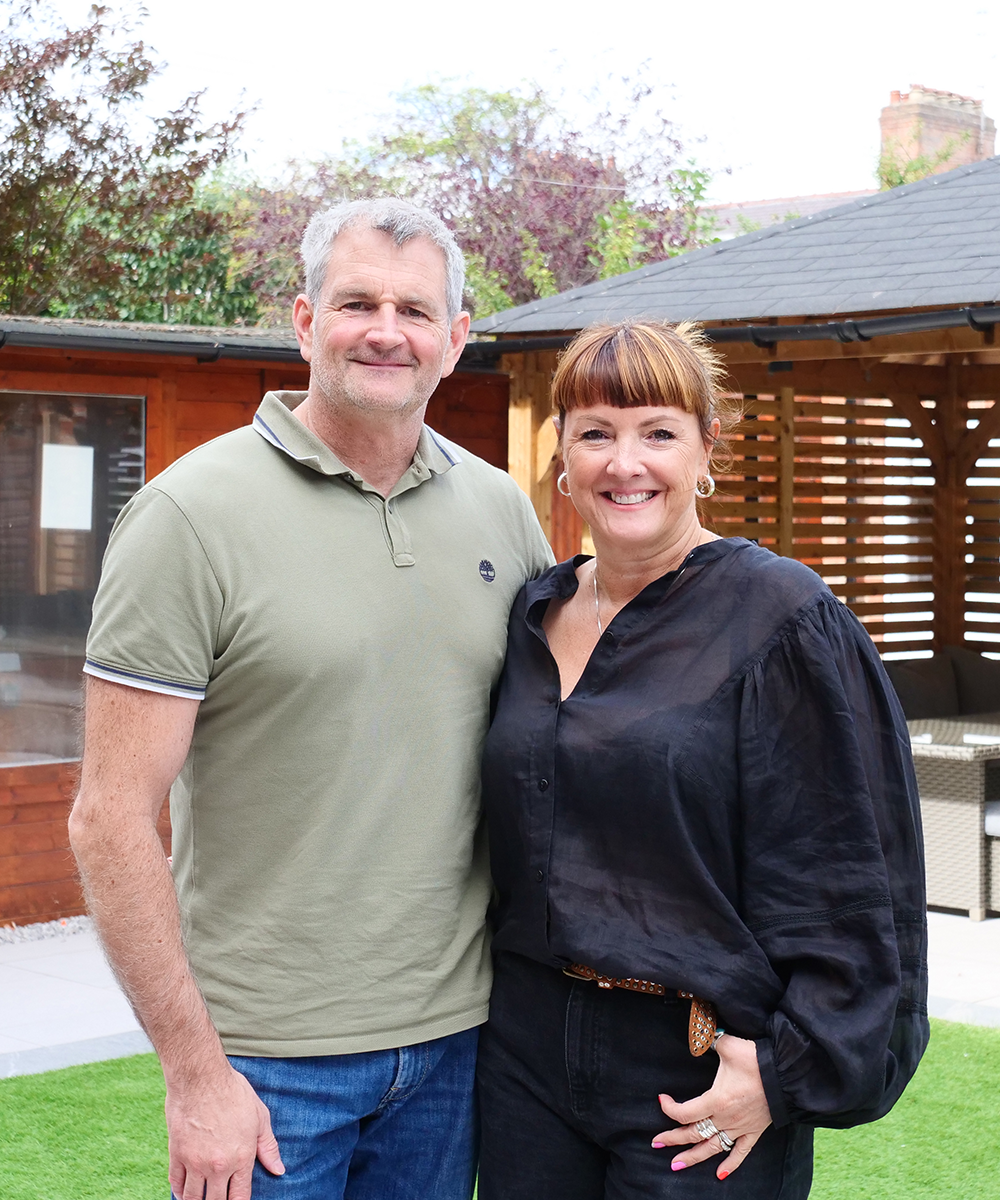 Man and woman smiling, posing in a backyard. Man wears olive green polo and jeans. Woman wears black top and pants.