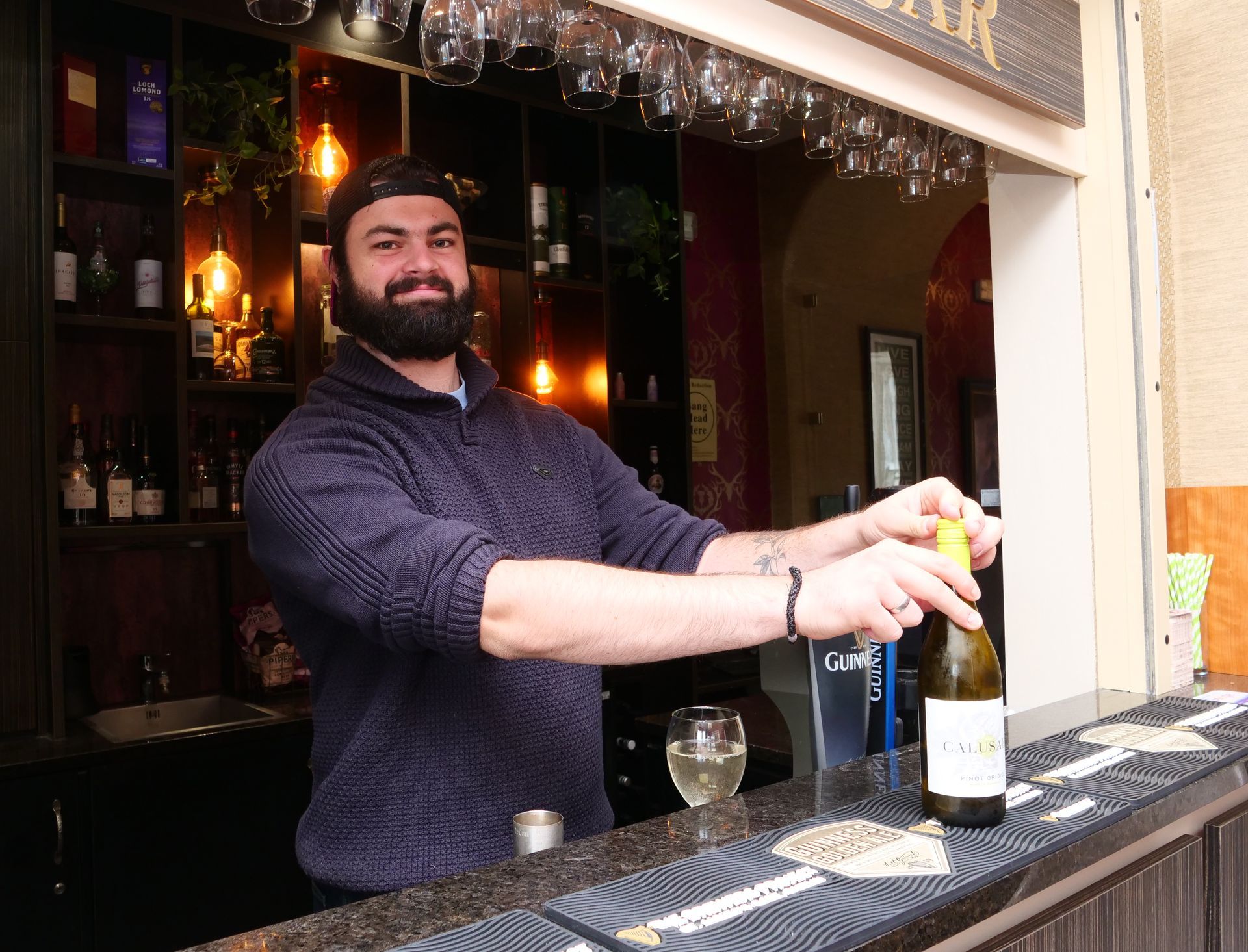 Bartender with a beard opens a wine bottle at a bar. The setting is outdoors, with wine glasses hanging above.