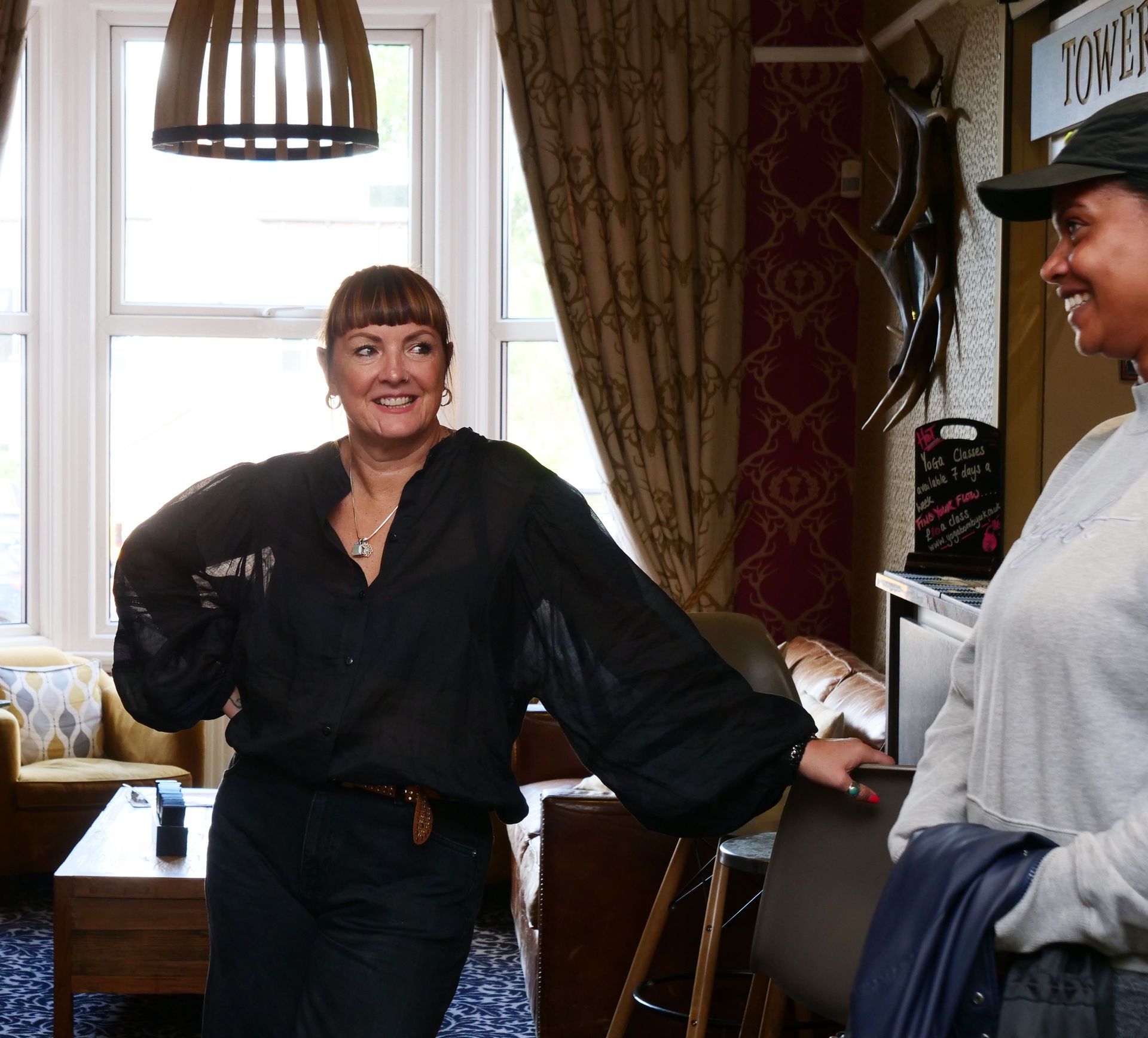 Two women in a pub, smiling. One wears black, the other a gray hoodie. Interior with bar and windows.