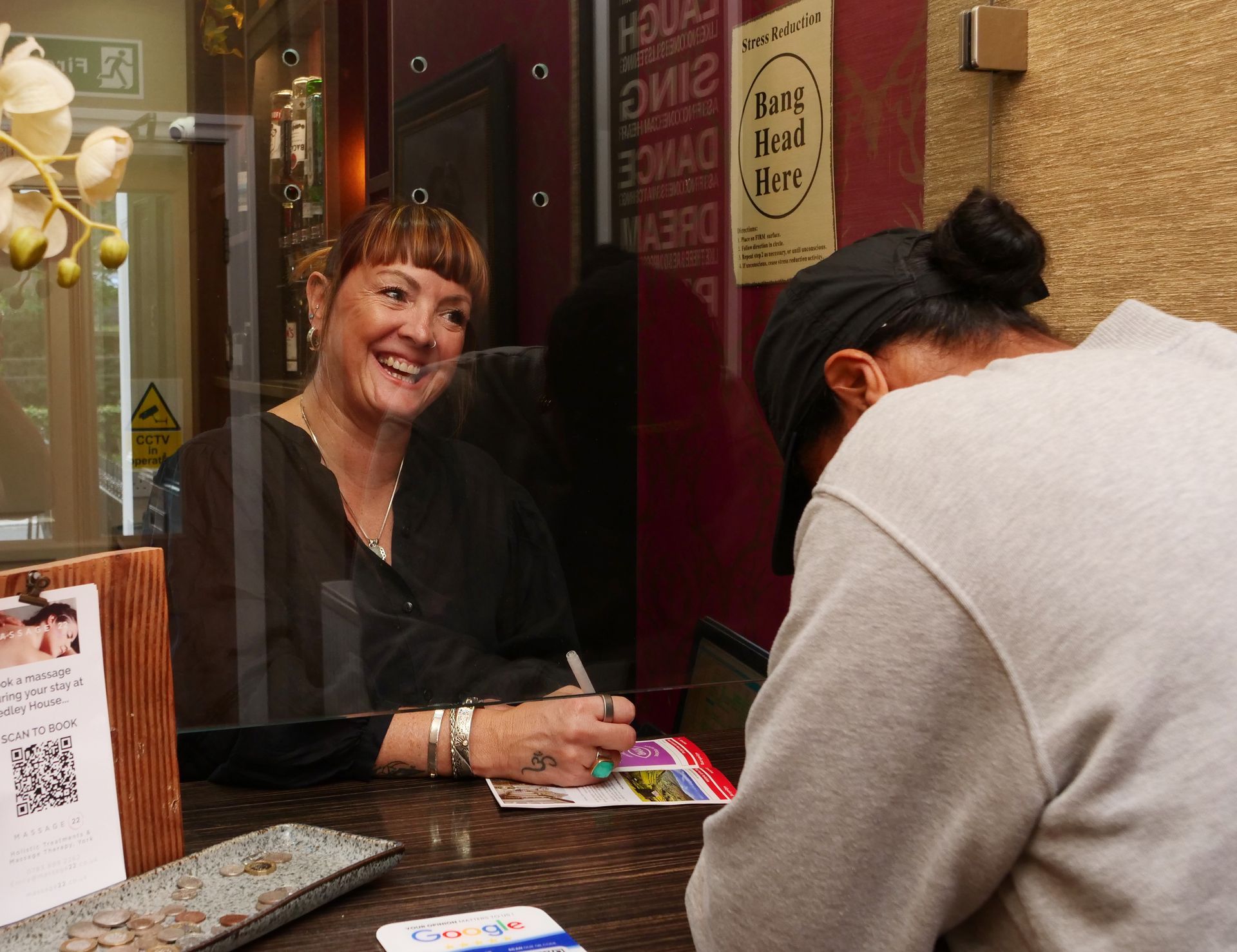 Woman smiles, signing something for a person at a counter. Clear barrier separates them.