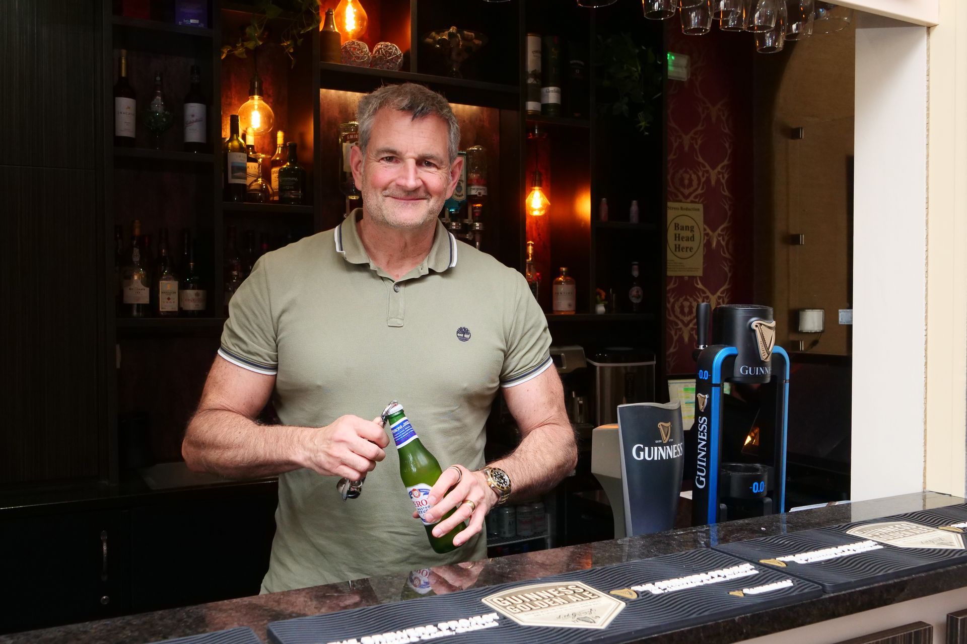 Man opening a beer bottle behind a hotel bar. He's smiling, wearing a green shirt. Interior with dark wood and lights.