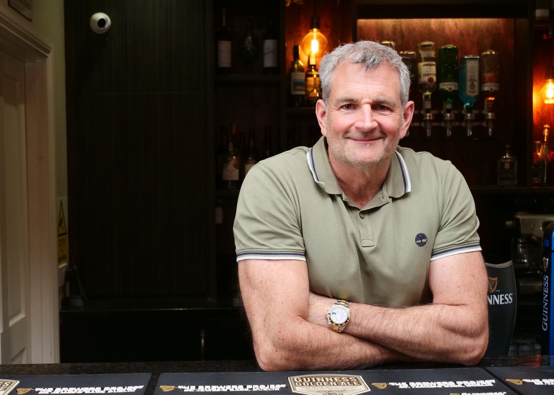 Man with gray hair, arms crossed, smiling at a bar. He wears a green polo shirt.