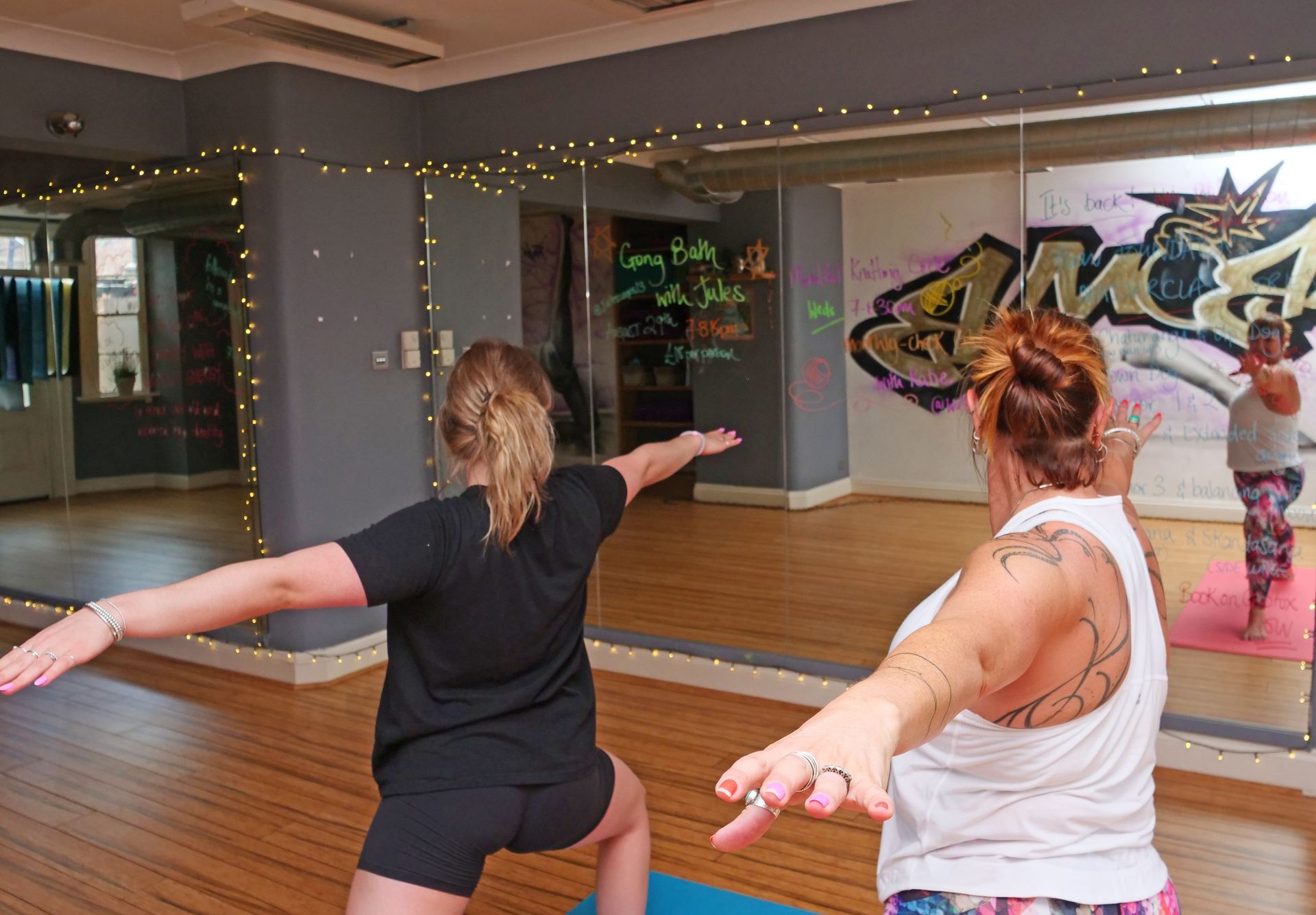 Two women in yoga pose, arms outstretched, facing a mirror in a studio.