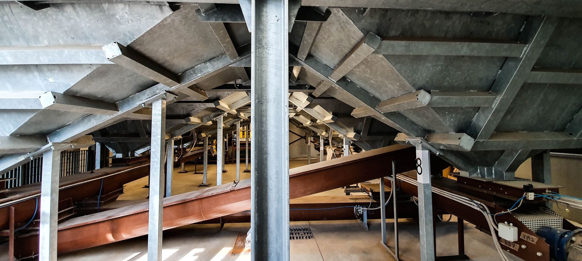 Struttura in acciaio: travi, colonne e un sistema di nastri trasportatori. Dominano i toni del grigio, ambiente industriale.