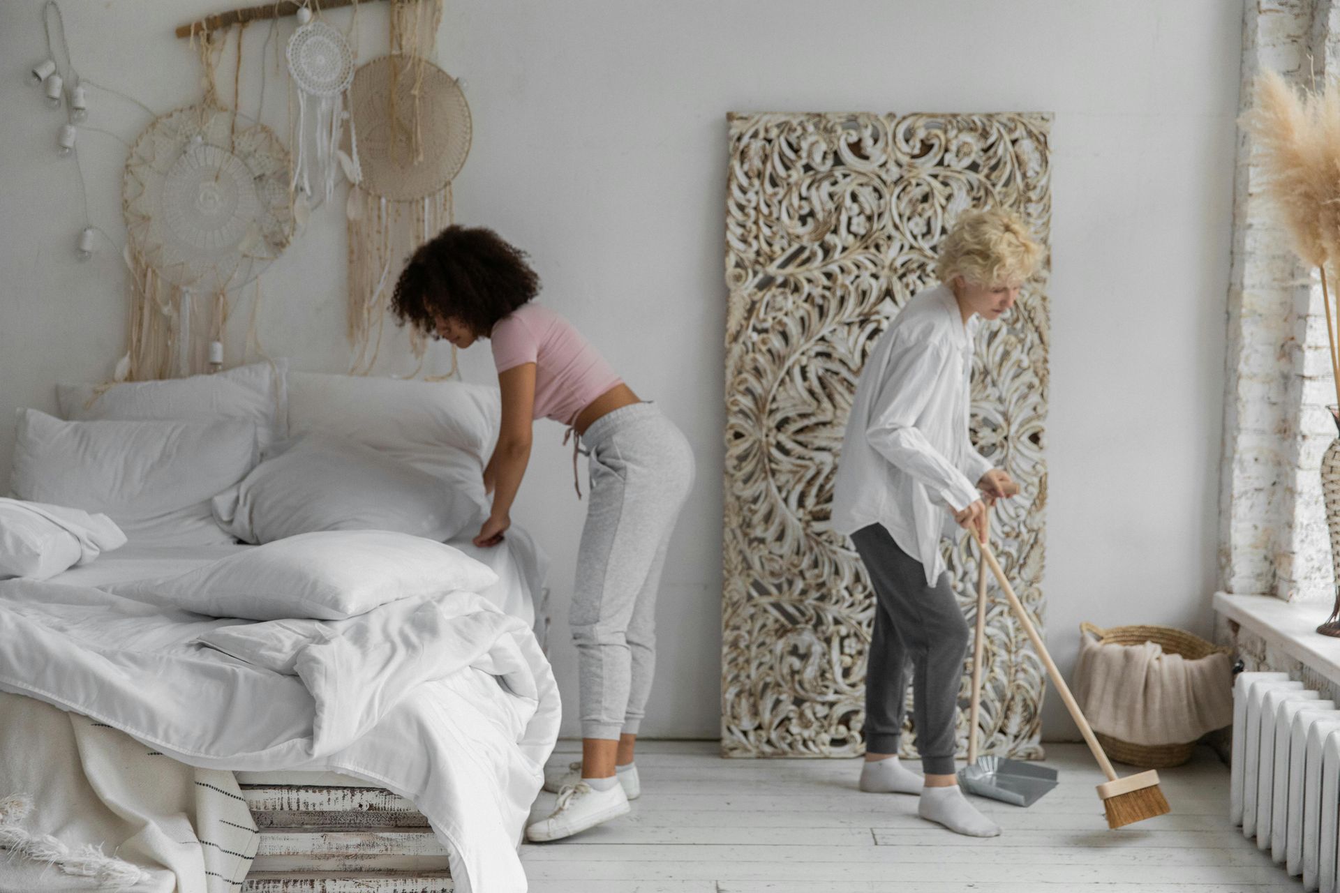 Woman making bed as another sweeps white floor. Room has white walls, decorations.