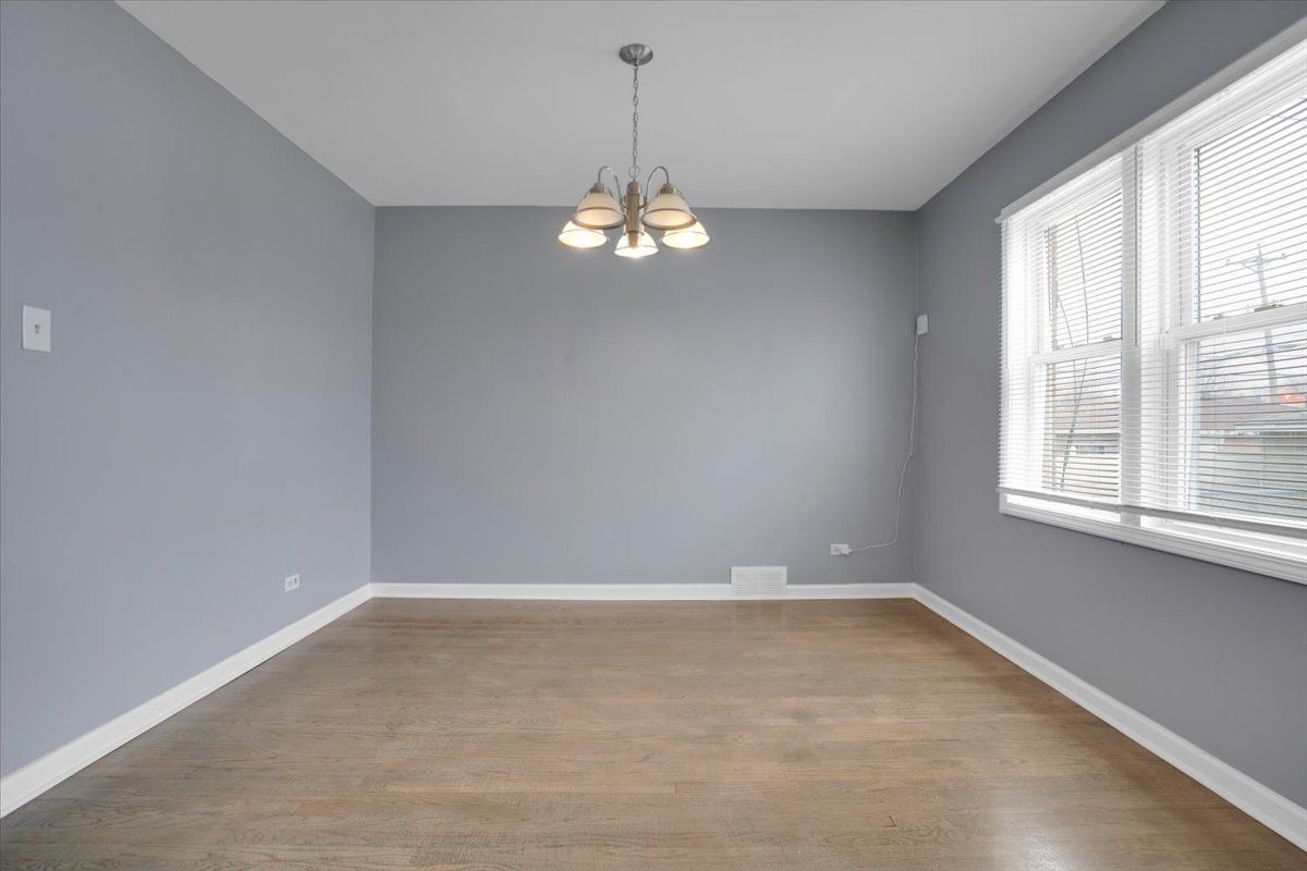Empty dining room with gray walls, wood floor, chandelier, and window with blinds.