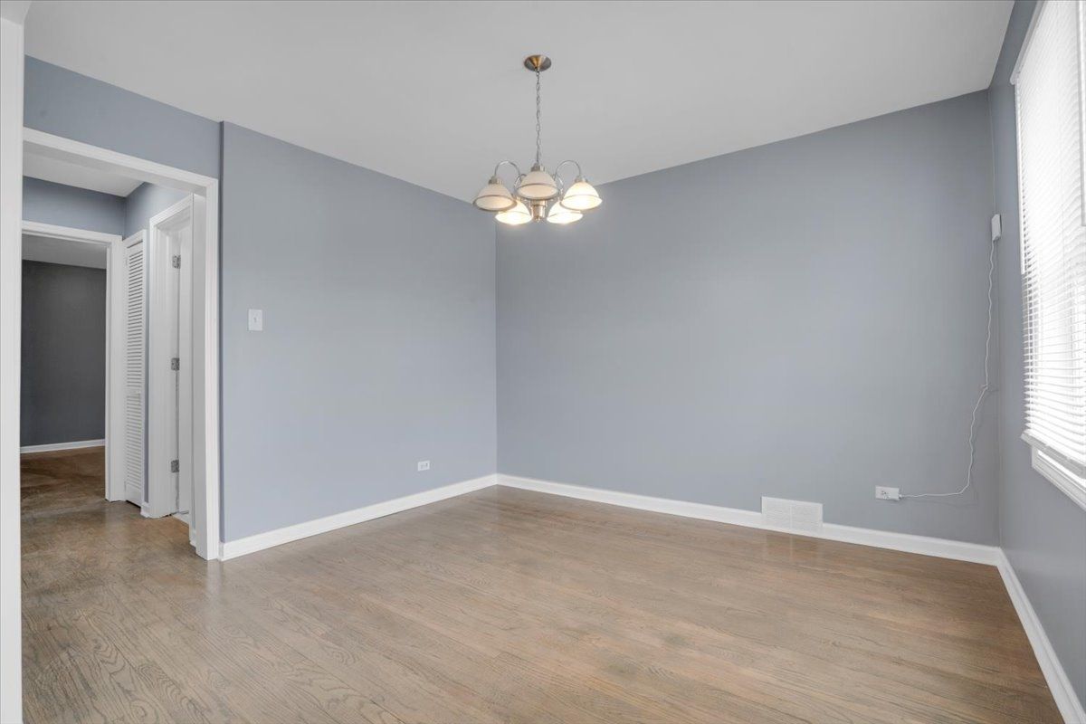 Empty room with gray walls, wood floor, and a chandelier. Light enters from a window.
