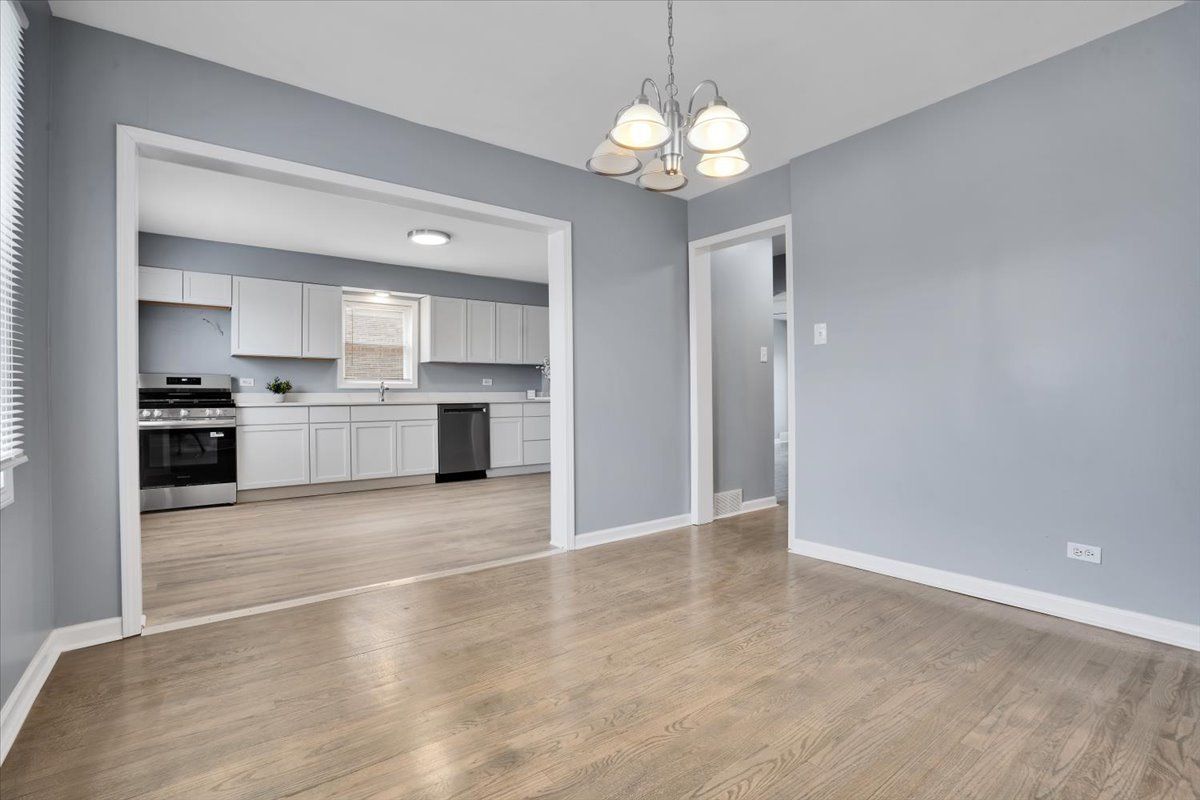 Empty dining room with light blue walls, light wood floor, and view into a white kitchen.