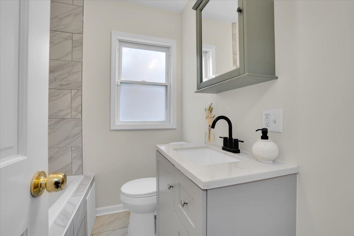 Small bathroom with gray vanity, toilet, and window. White walls, marble-look tile, and a black faucet.