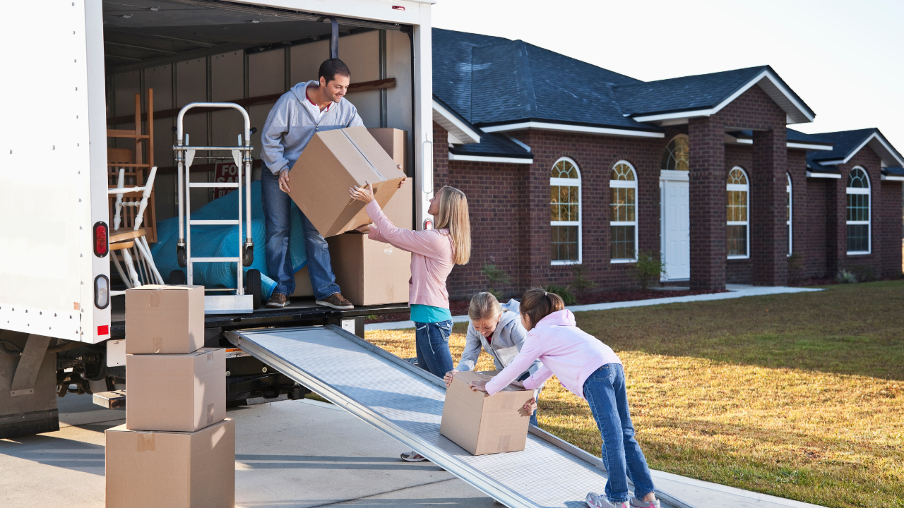 Family unloading moving truck in front of new home; father hands boxes to children, sunny day.