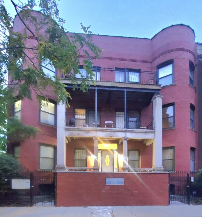 Red brick three-story building with a porch and curved tower. Lit entryway, with trees in the foreground.