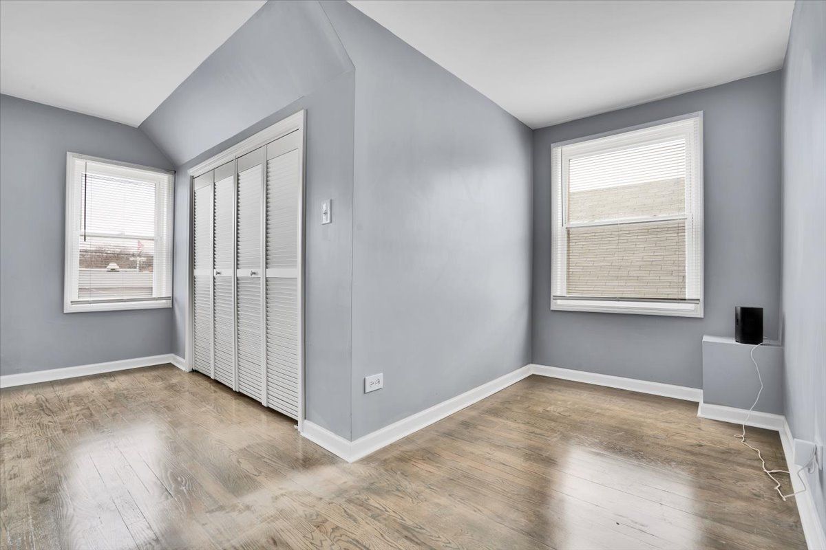Empty bedroom with gray walls, white trim, wooden floor, and a closet. Two windows.