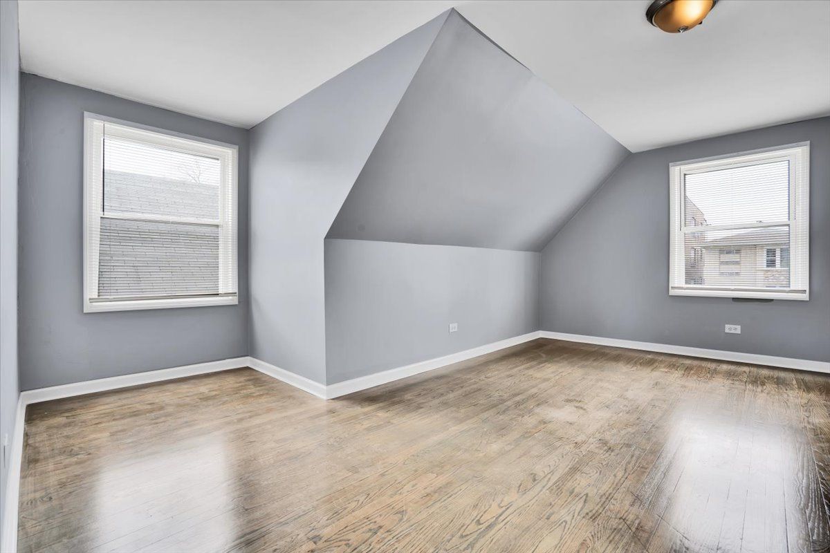 Empty gray room with angled ceiling and two windows; wood floor.