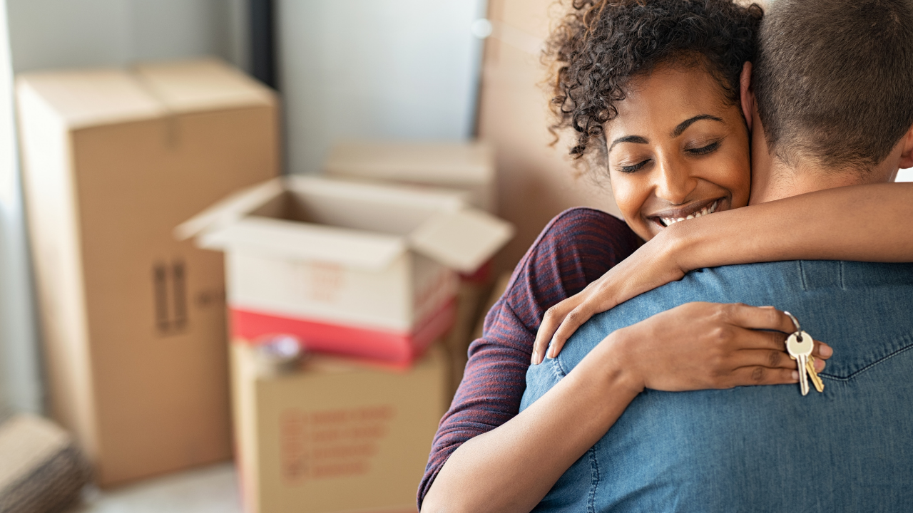 Woman hugging a man, smiling, holding keys, boxes in background, moving into a new home.