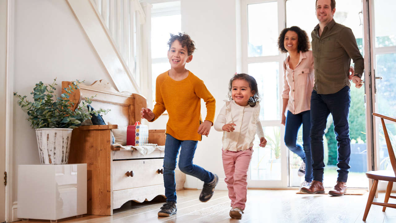Two children run joyfully into a bright home entryway as two adults follow them through an open door.