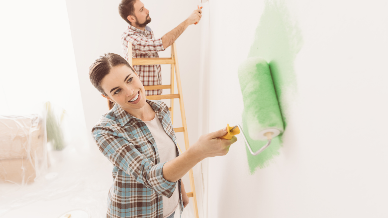 Woman and man painting a wall green. Woman smiling, man on ladder. Bright, indoor setting.
