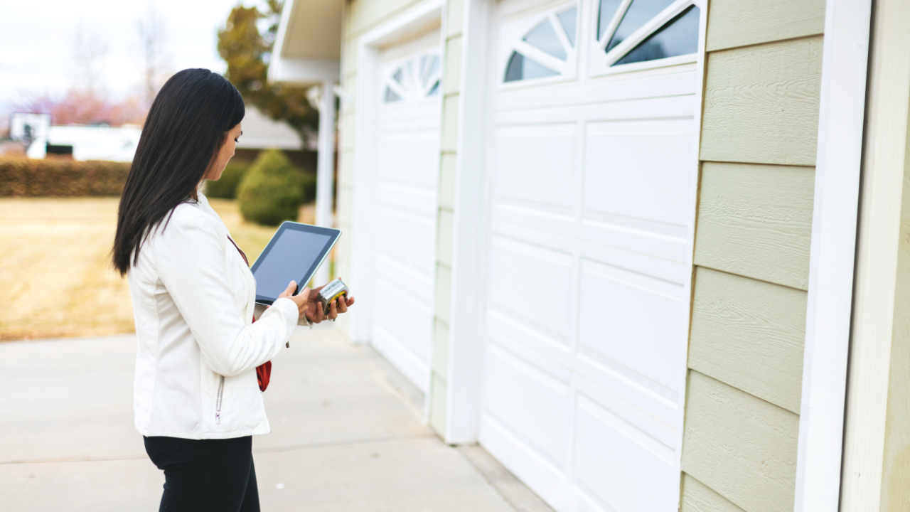 Woman in white jacket, holding a tablet, stands near a white garage door in a suburban neighborhood.