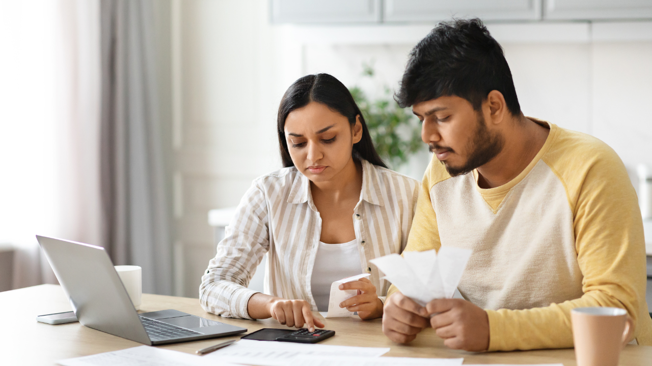 Couple reviews finances together, using calculator and laptop in a home setting.