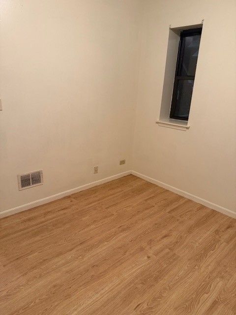 Empty room with wood-look flooring, cream walls, small window with dark interior, and a heating vent.