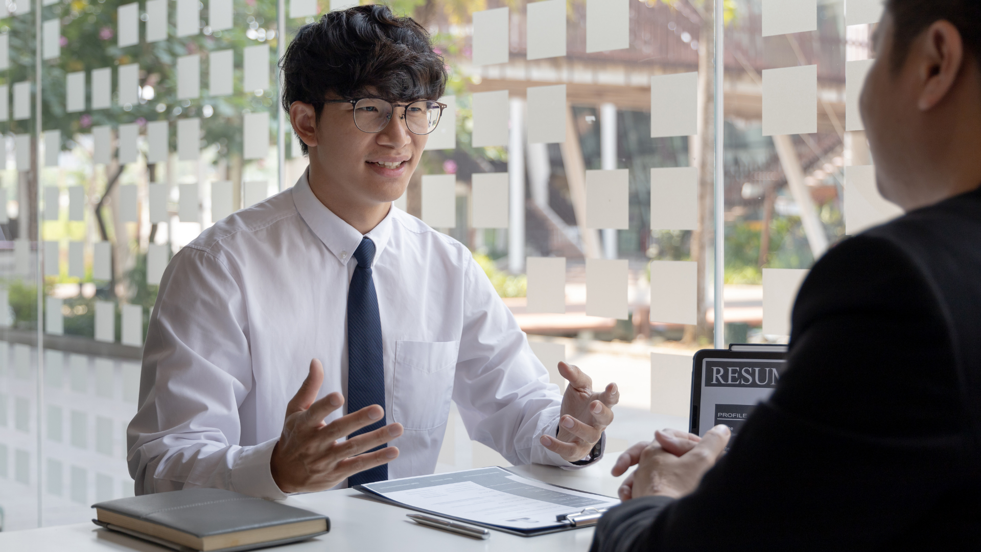 Man in white shirt and tie speaks to another man at a desk in a well-lit room.