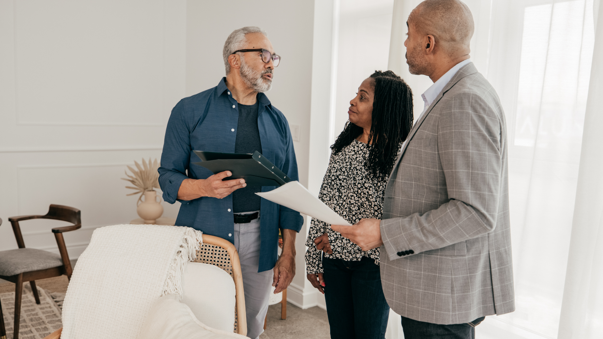 Real estate agent shows home to older couple in a bright room, all wearing business casual.