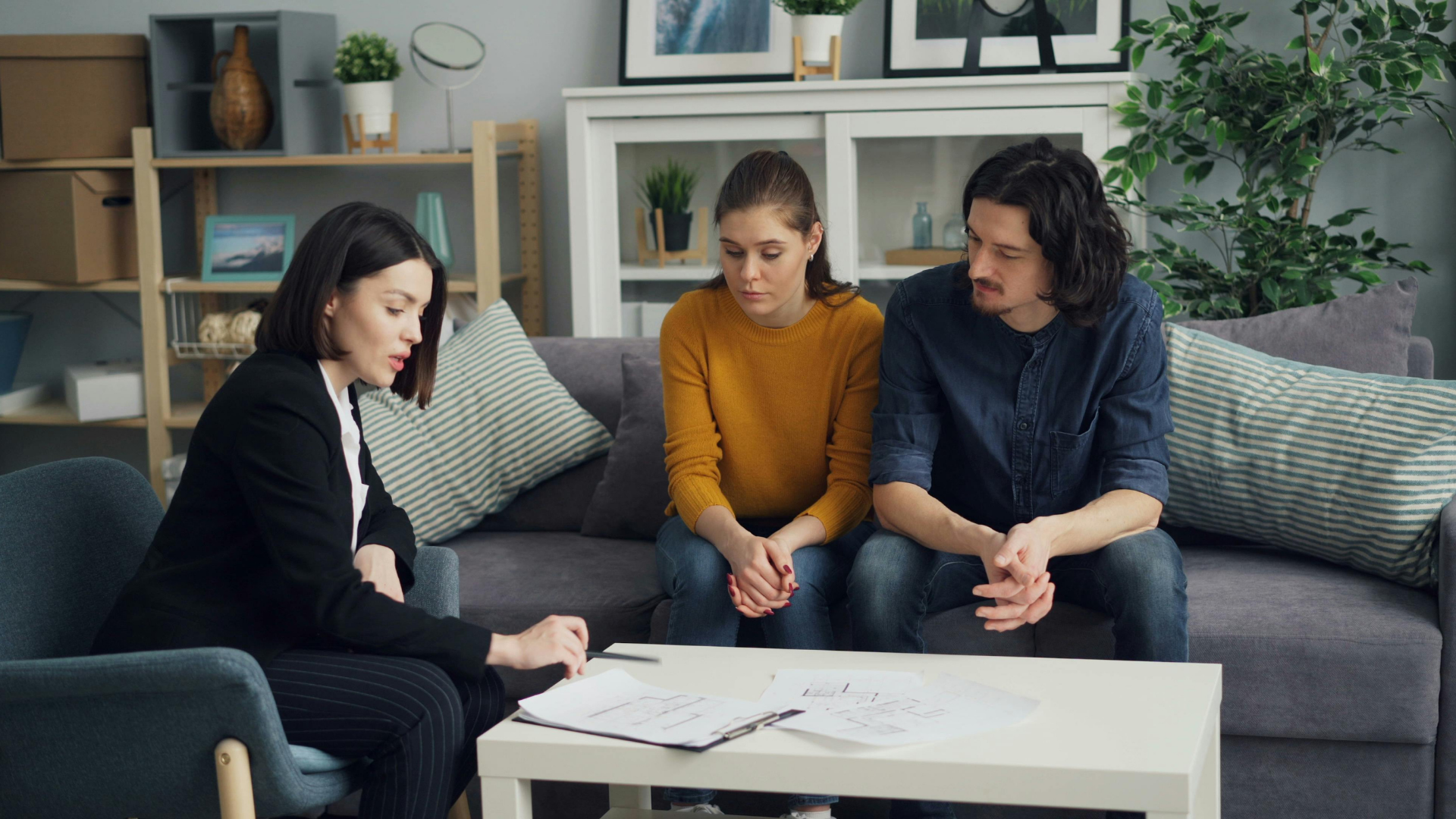 A woman in a blazer reviews papers with a concerned couple on a couch in a living room.