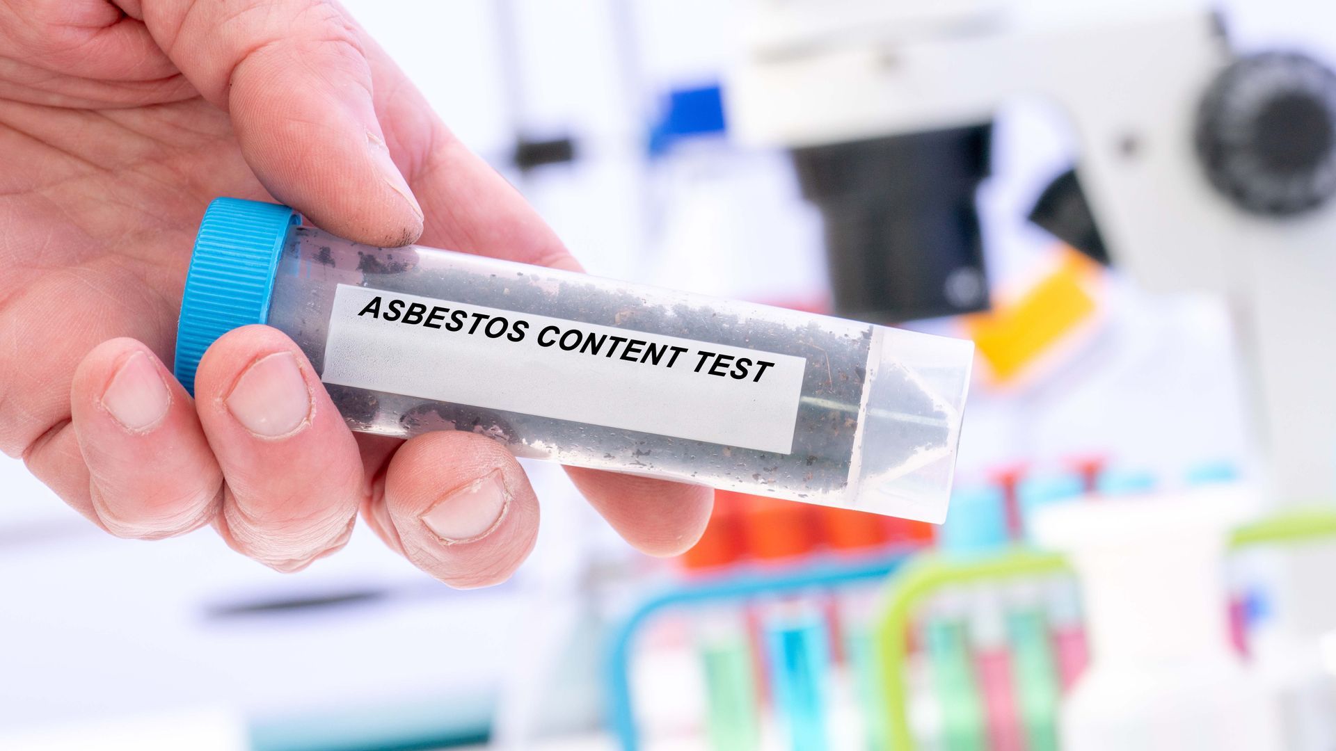 Hand holding a labeled test tube in a lab with colorful samples blurred in the background