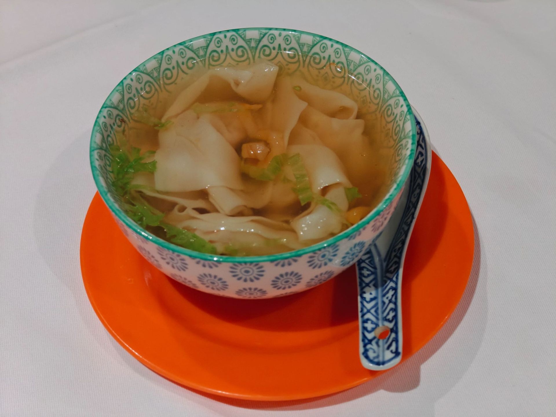 Una ciotola di zuppa wonton con un cucchiaio di ceramica abbinato è adagiata su un piatto arancione su sfondo bianco.