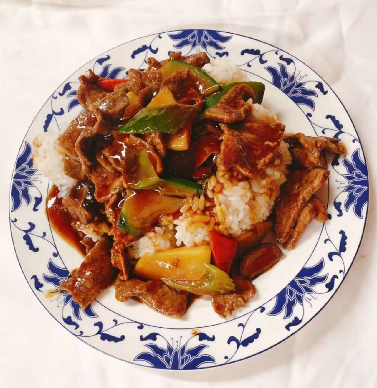 Un piatto di manzo e verdure saltate in padella, servito su riso bianco e presentato su un piatto bianco con un bordo floreale blu.