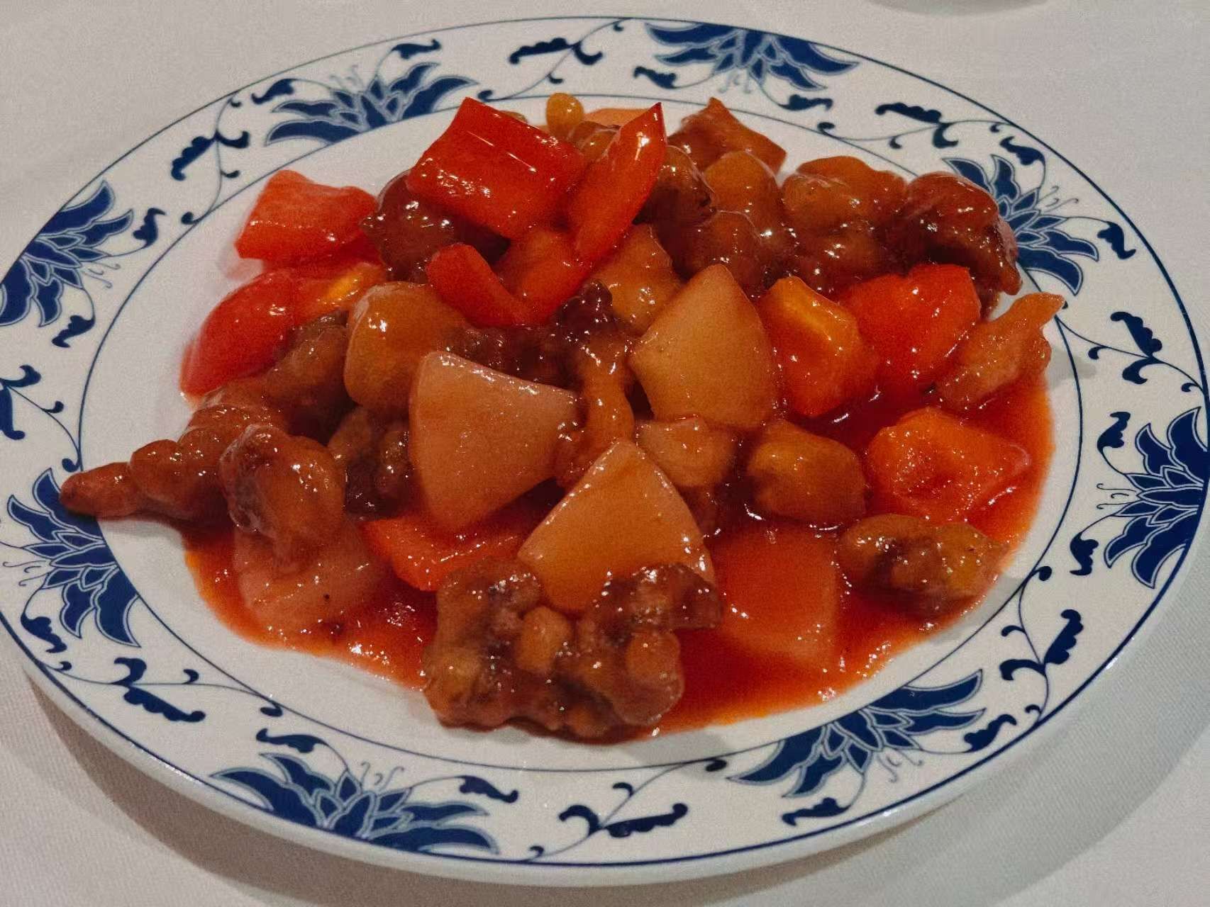 Un piatto di maiale in agrodolce con pezzi di carne, ananas e peperoni rossi in una salsa rossa lucida.