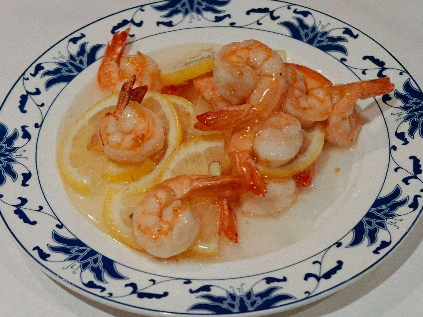 Gamberi cotti serviti su un piatto con sottili fette di limone e una salsa leggera, su un bordo decorato con motivi blu e bianchi.