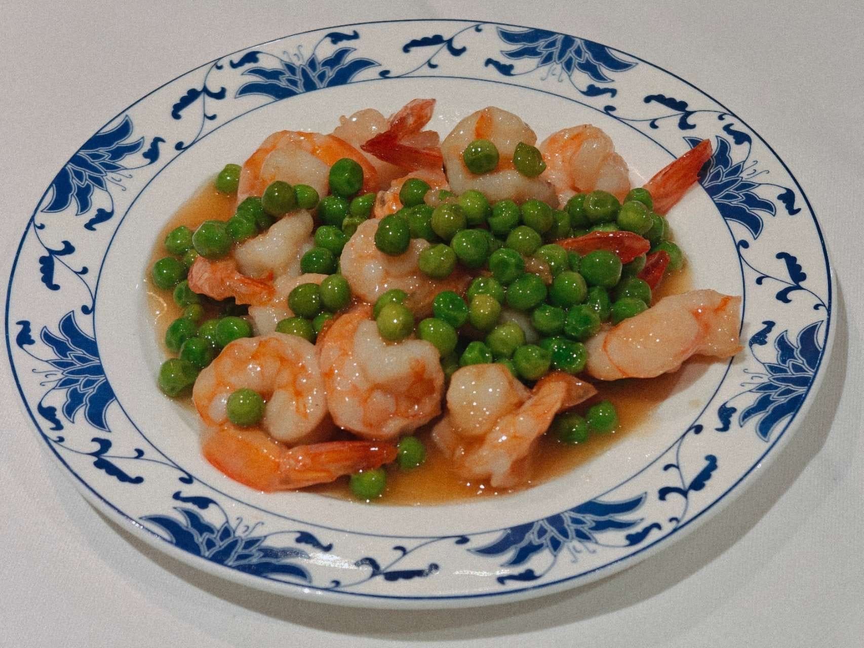 Un piatto di gamberi con piselli in una salsa leggera e brunastra, servito su un piatto bianco con il bordo decorato con fiori blu.