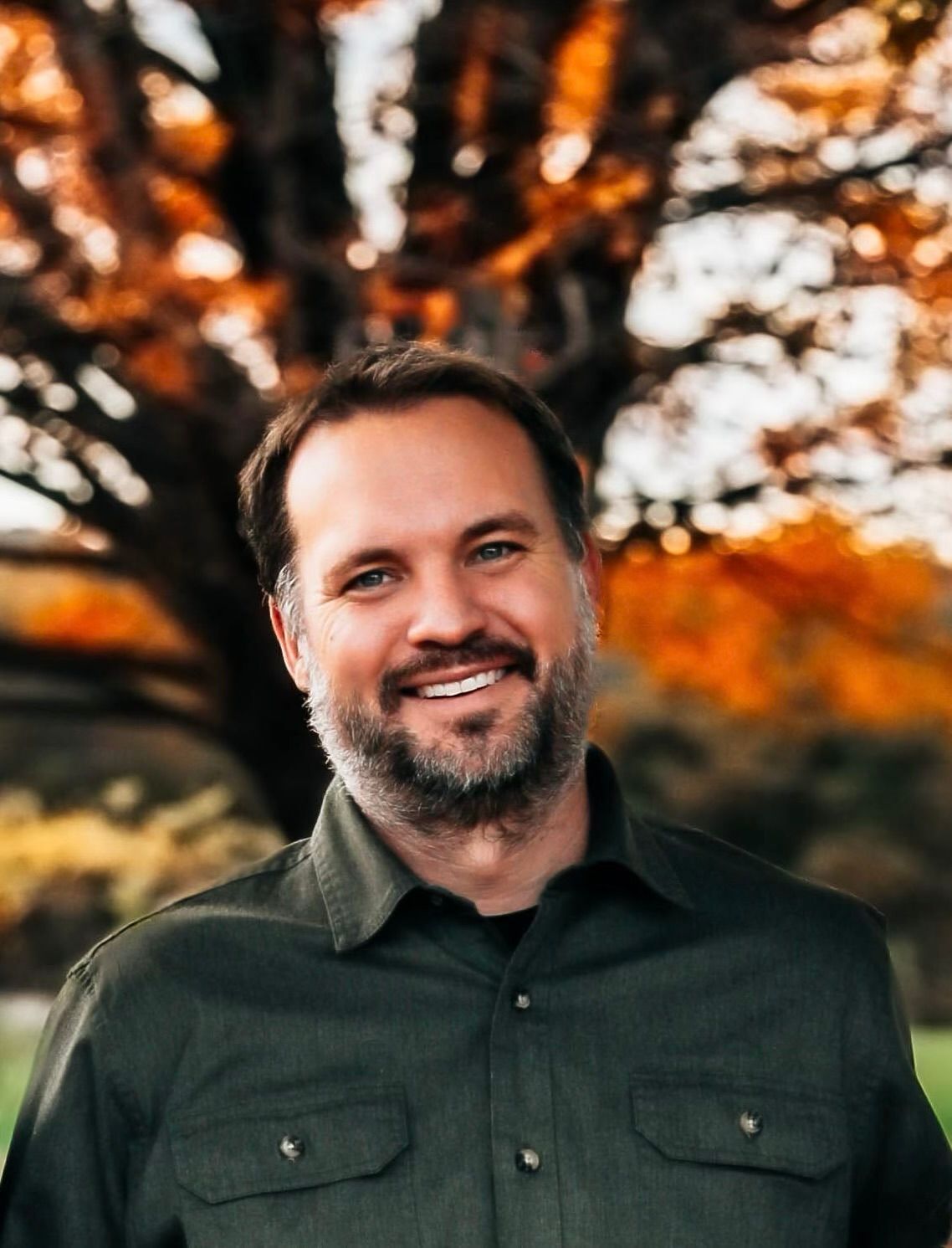 A man with a beard is smiling in front of a tree.