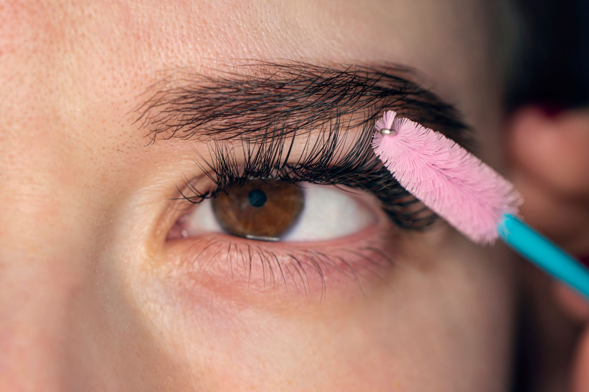 A woman is applying mascara to her eyelashes with a pink brush.