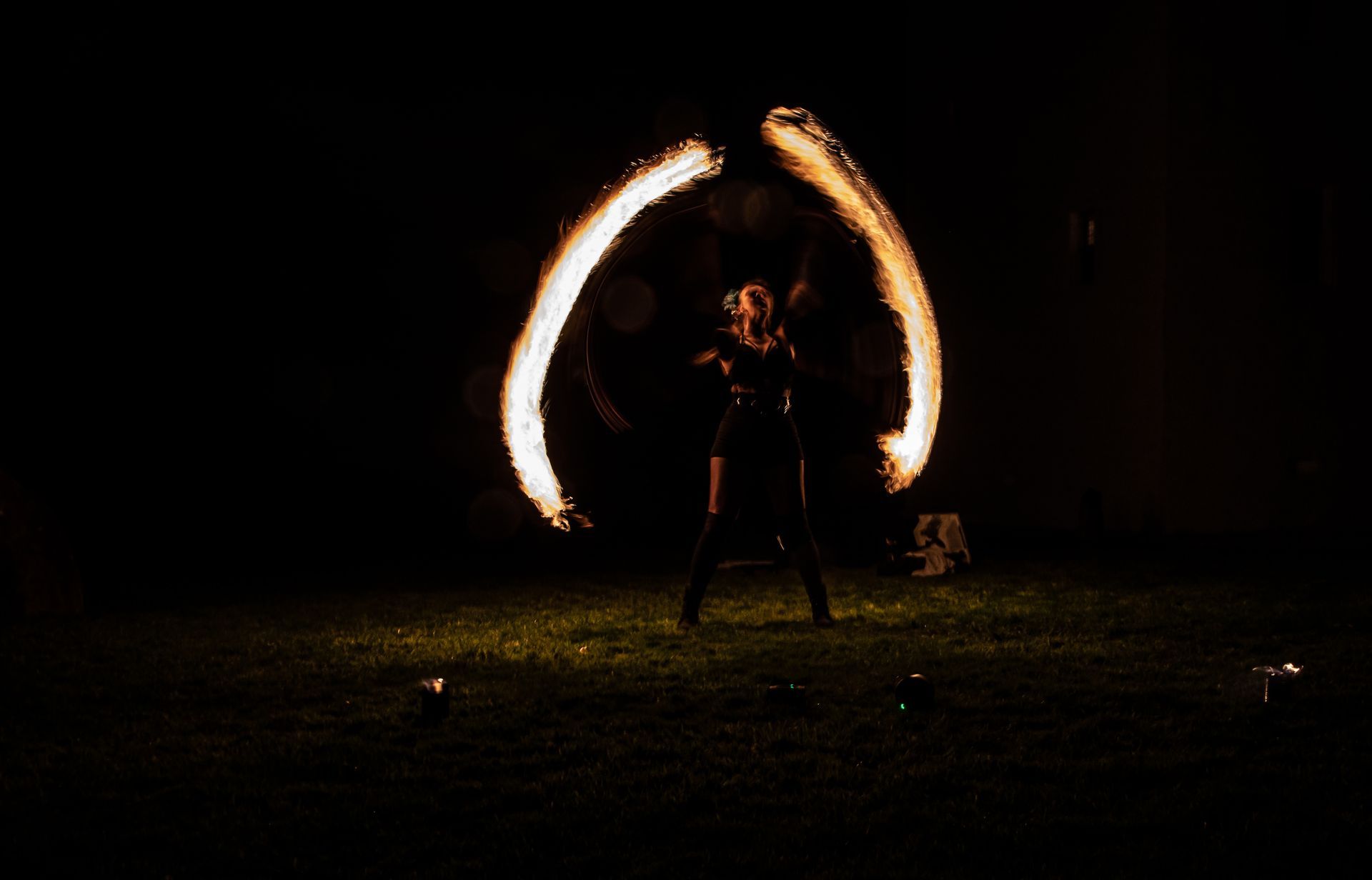 a colour image of Mia Meow stood on some grass in the dark, you can barely make out the performer but there are two huge columns of light each side of her created by the fire trails by Lorna Newman Photography