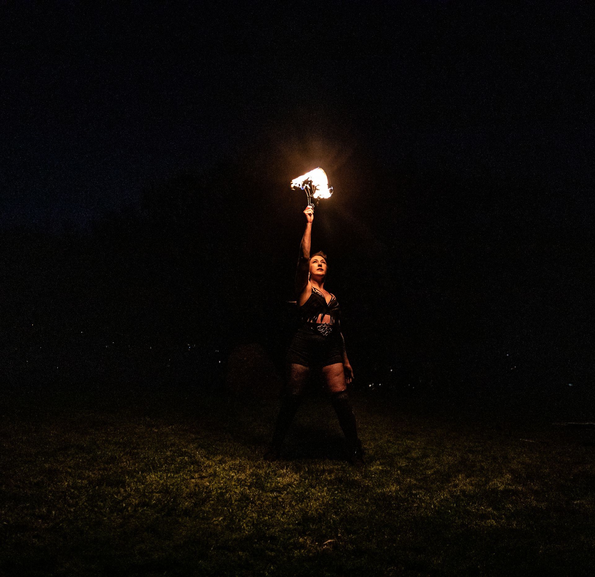 a colour image of Mia Meow stood on some grass in the dark, her hand is upstretched holding a baton of flames above her head, her other hand by her side in a power stance by Lorna Newman Photography