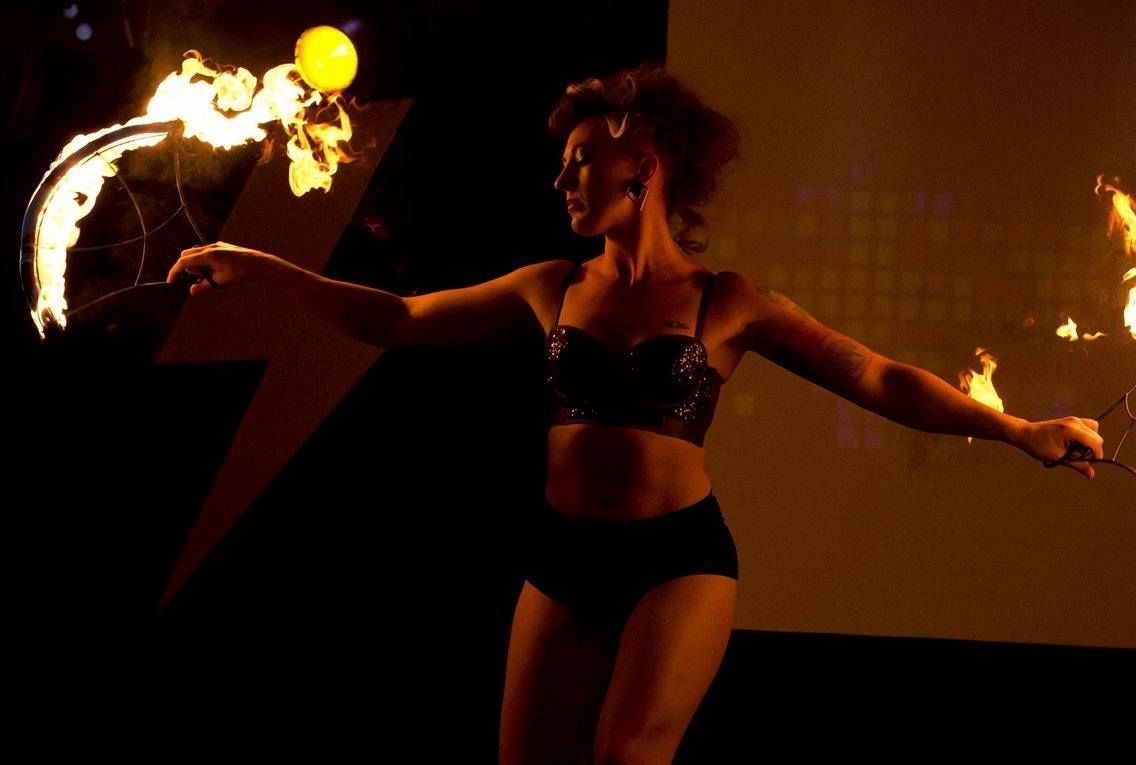 a colour image of Mia Meow with arms outstretched to either side, she is dancing on stage lit by a soft warm light and has a fire fan in each hand which are giving soft trails by Jamie McFadden