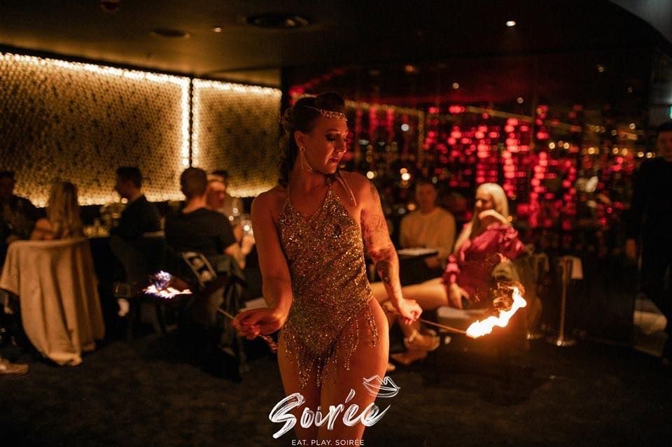 Colour photo of Mia Meow in a cabaret venue with audience seated around, she is sashaying through the audience with lit fire wands in each hand by soiree / Devil May Care Agency