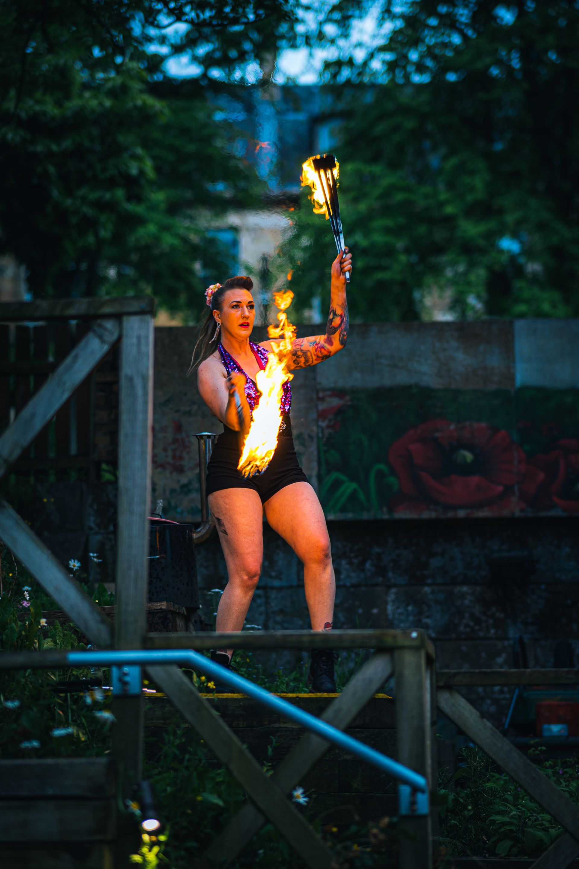 A colour image of Mia Meow stood on a platform surrounded by greenery, she has a slightly sideward stance with two batons of fire in each hand, one is mid way in front of her the other midway up as she dances by Andres McNeill