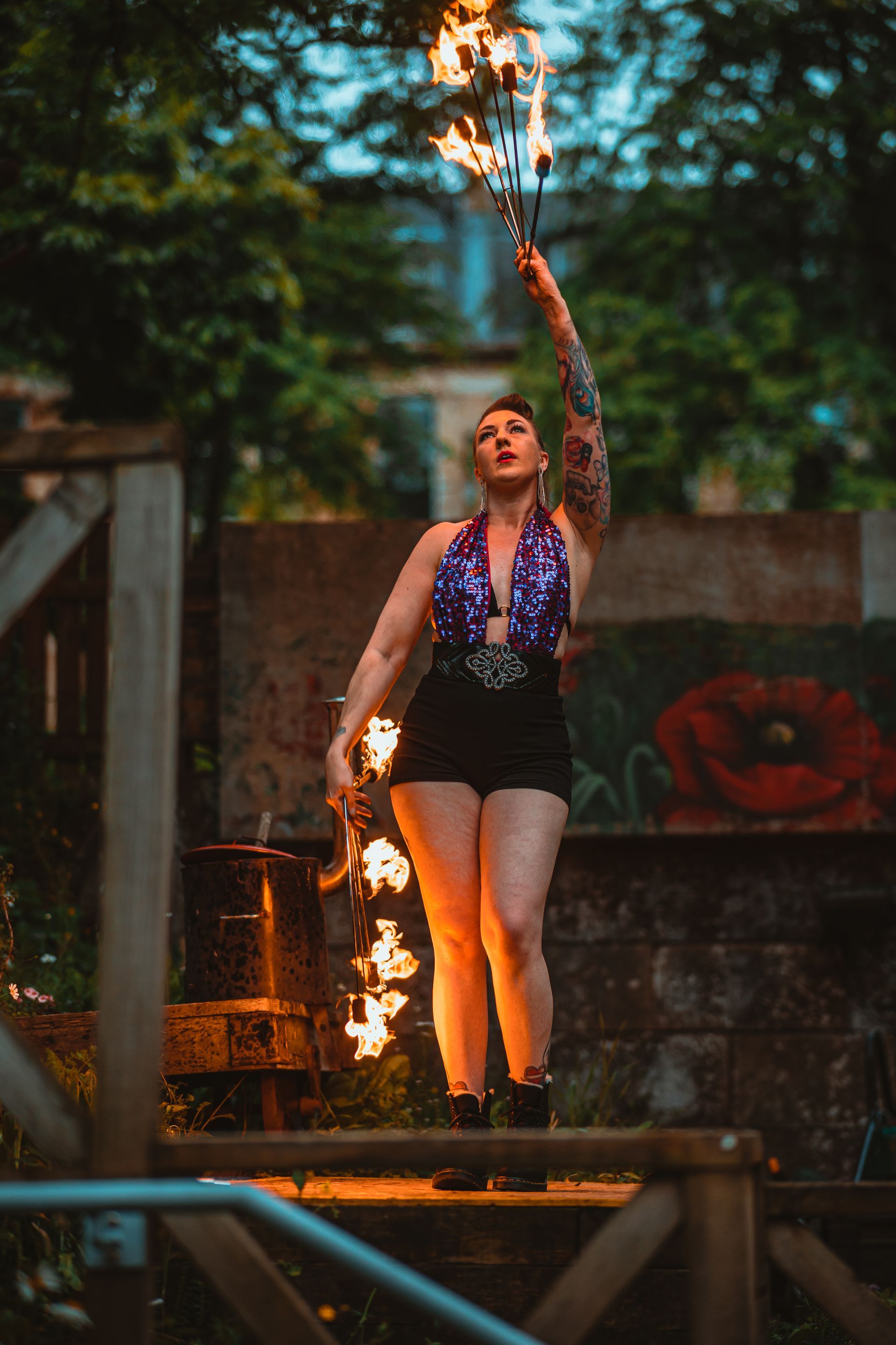 A colour image of Mia Meow stood o a platform surrounded by greenery, she has a fire fan in each hand with one arm up above her head and the other down by her side mid-dance by Andres McNeill
