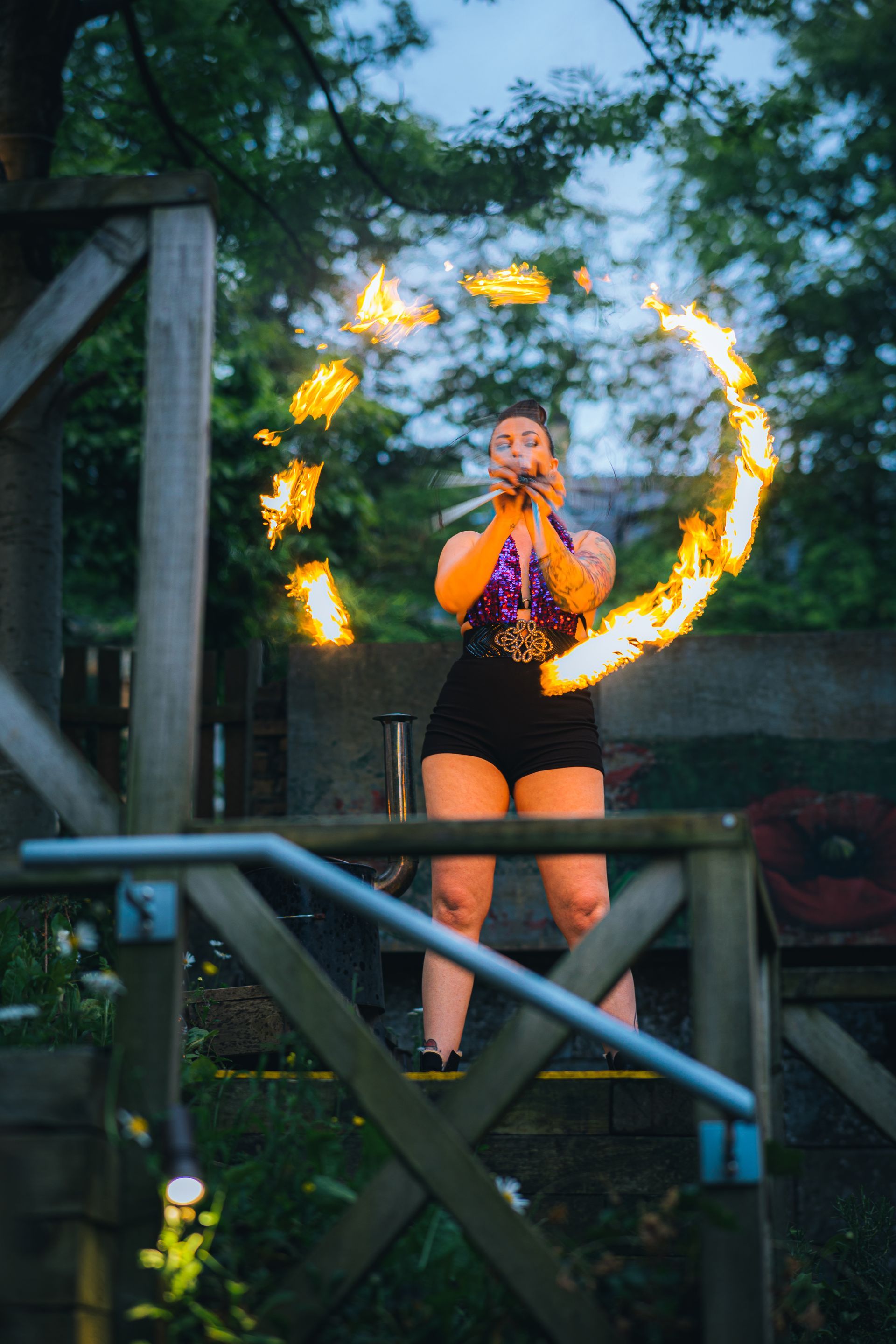 Colour image of Mia Meow stood on a platform surrounded by trees, she has her hands together arms outstretched in front of her with a ring of fire created by fire fans in front of her by Andres McNeill