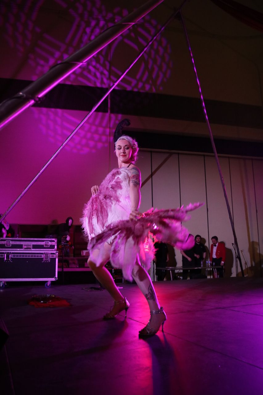 A colour photo of Mia Meow lit in pinks an purples on stage with two feather fans in pink and white by Amy Shipway