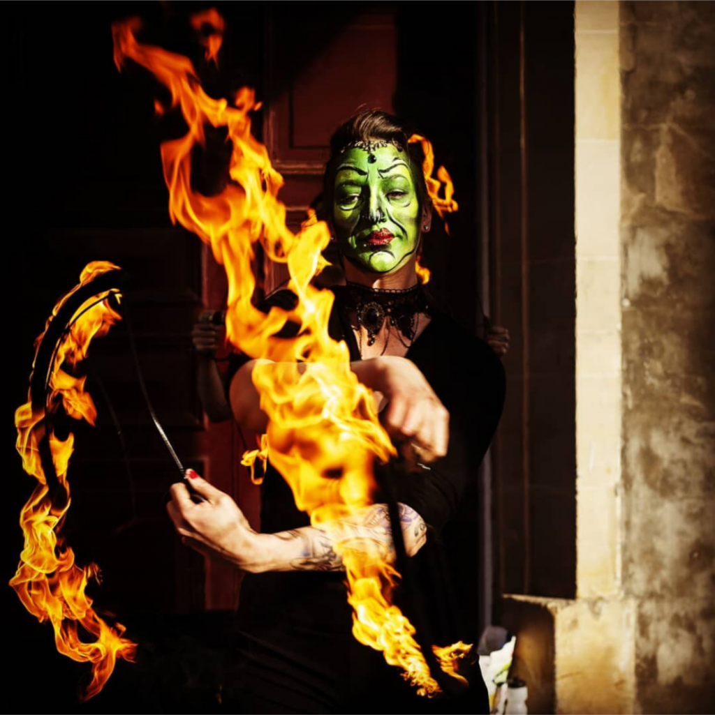 a colour photo of Mia Meow with large flame trails in front of her dressed as a witch with green face paint by Kerrin Carr Photography